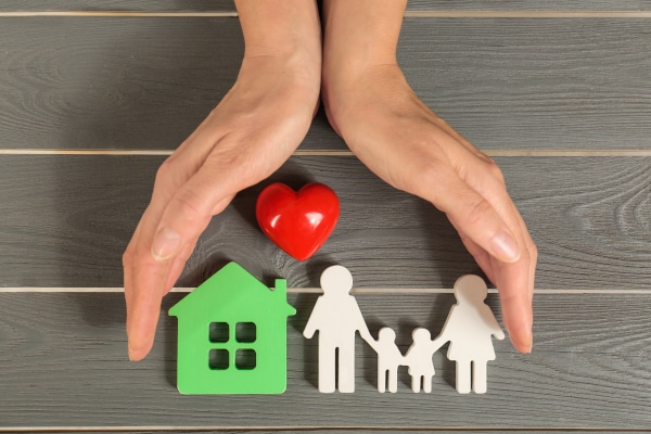 Image of a pair of hands sheltering a model house and a wooden family cutouts with a red heart