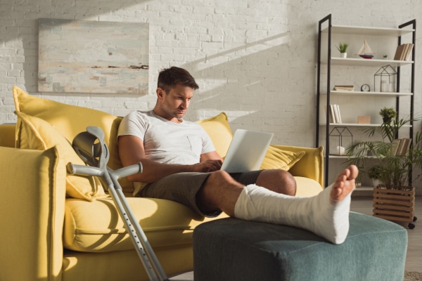 Young, fit man with a broken leg sitting on a sofa with a laptop and a pair of crutches leaning against the sofa