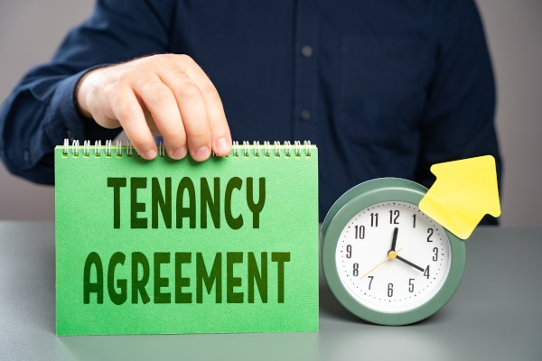 Image of a hand holding a greed backed notebook with teh words Tenancy Agreement printed on the front with a clock and a paper yellow house stuck on the front