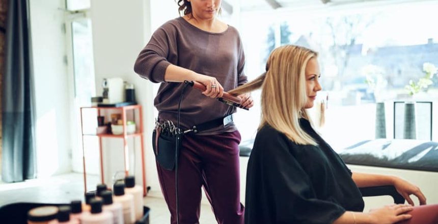Young hairstylist using a straightener on the hair a female client sitting in a chair while working in her salon